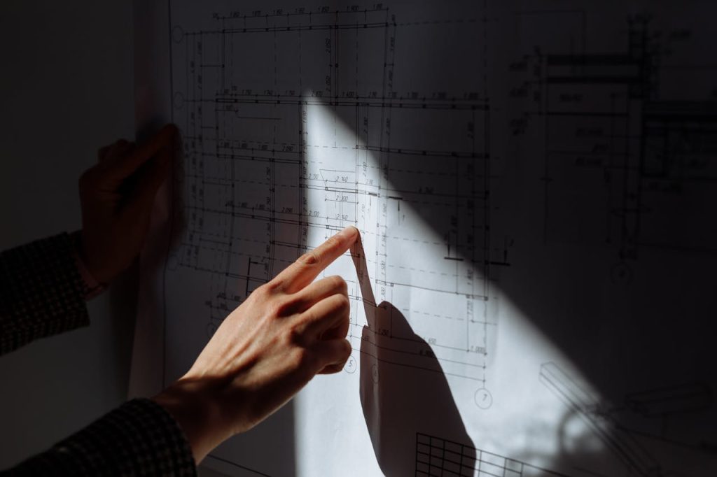 finger-pointing-the-floor-plan-8470847 A detailed examination of construction blueprints under dramatic lighting.